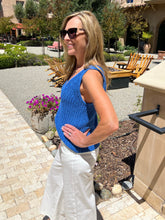Woman in a blue sleeveless top and white pants standing outdoors with garden and building in the background