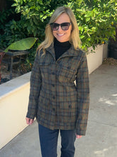 A woman wearing a green and brown plaid button-up jacket over a black turtleneck and dark jeans, standing outdoors on a concrete walkway with green foliage and a beige wall in the background.