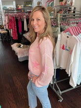 Woman in a pink sweater and jeans standing in a clothing store with racks of clothes in the background.