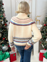 Woman wearing a cream turtle neck sweater with a colorful pattern showing the back view in a festive setting with a decorated Christmas tree.