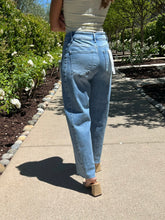 Person wearing light blue jeans and a white top walking on a paved path with greenery in the background