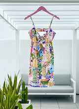 Colorful patterned dress hanging on a hanger against a white wall with a plant in the foreground.