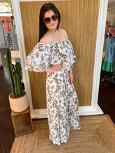 Woman in a floral off-shoulder top with matching pant, standing against a wooden panel wall.