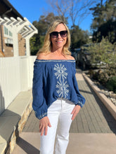Woman wearing a blue embroidered blouse and white pants standing on a wooden deck.