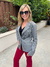 A woman wearing a gray patterned blazer, black top, and red pants stands outdoors in a garden setting.