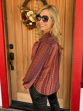 A woman wearing a long-sleeve patterned blouse with orange, red, and brown tones, paired with black pants, standing in front of a wooden door with a fall wreath.