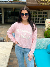 Woman wearing a pink sweatshirt with 'mermaid vibes' text, standing outdoors.