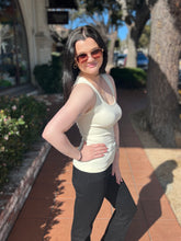 Woman wearing a white tank top and black pants standing on a sidewalk with trees and buildings in the background.