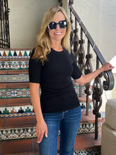 A woman with blonde hair wearing a black short-sleeve crewneck top and blue jeans, standing on patterned tiled stairs next to a black wrought-iron railing.