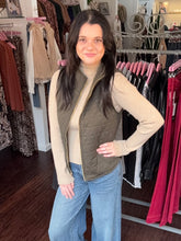 A woman modeling a sleeveless olive green quilted vest over a beige turtleneck sweater and blue jeans, standing inside a clothing boutique with racks of garments in the background.