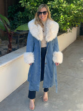 A woman wearing a long light blue denim coat with a large white faux-fur collar and matching cuffs, standing outdoors on a concrete walkway with green plants in the background.