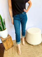 Person wearing blue jeans and a black top standing in a room with a plant and ottoman.