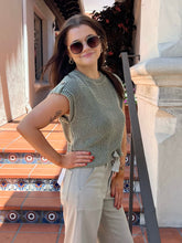 A woman wearing a ribbed sage green sleeveless top and beige pants, standing on tiled outdoor steps next to a white stucco building.