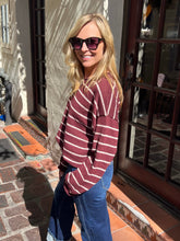 A woman wearing a maroon and cream striped long-sleeve knit top and blue jeans, standing outdoors in front of a building with glass doors and stone steps.