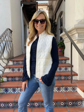 A woman wearing a cream quilted zip-front puffer vest over a navy long-sleeve top and blue jeans, standing on outdoor tiled steps in front of a building.