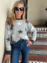 A woman wearing a light gray knit sweater with dark gray and white star patterns, paired with dark blue jeans and black sunglasses, standing in front of a tiled staircase.