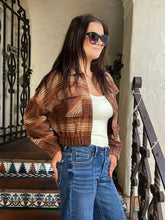 A woman wearing a brown and tan plaid cropped jacket, a white tank top, and blue jeans, standing on an outdoor staircase with a stucco wall and black wrought iron railing behind her.