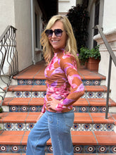 A woman wearing a pink and orange abstract patterned long-sleeve mock-neck top and blue jeans, standing on terracotta tiled outdoor stairs.
