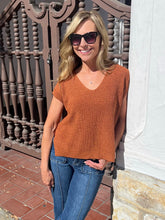A woman wearing a rust-orange ribbed knit V-neck short-sleeve sweater and blue jeans, standing outdoors in front of a decorative brown metal gate and a white wall.