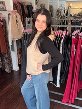 A woman posing in a clothing boutique wearing a beige quilted vest over a black long-sleeve top and blue jeans, with racks of clothes visible in the background.
