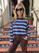 A woman wearing a brown and light blue striped knit sweater and brown pants, standing on outdoor tiled steps in front of a building.