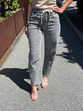 A person outdoors wearing gray leopard-print jeans with a drawstring waistband, paired with a beige top and nude heeled sandals on a paved walkway.