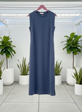Blue sleeveless midi dress hanging against a white background with plants below.