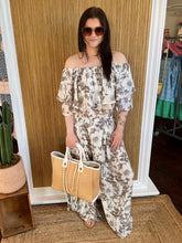 Woman in a floral off-shoulder top with matching pant holding a straw bag, standing against a wooden panel wall.