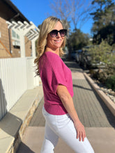 Woman in a magenta sweater and white pants standing outdoors on a sunny day.