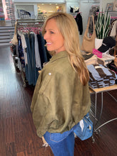 A woman with blonde hair wearing an olive green suede jacket and blue jeans, standing inside a clothing boutique.