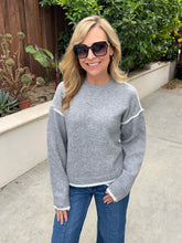 A woman wearing a light gray knit crewneck sweater with white contrast trim on the shoulders and cuffs, paired with blue jeans, standing outdoors on a concrete walkway with green plants and a wooden fence in the background.