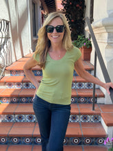 A woman wearing a light olive green short-sleeve V-neck t-shirt and dark blue jeans, posing on outdoor terracotta tiled stairs with decorative blue and white patterned tiles.