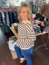 A woman with blonde hair smiling in a boutique, wearing a cream and brown striped knit sweater, blue jeans, and brown wedge sandals. Clothing racks and a display table are visible in the background.
