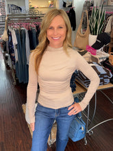 A smiling blonde woman wearing a beige mock neck long-sleeve top and blue jeans, standing inside a clothing boutique with racks of garments and tables in the background.