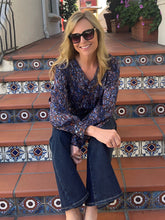 A woman wearing a long-sleeve blue and black patterned blouse and dark blue jeans sits on terracotta tiled steps with decorative tile borders.