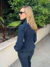 A woman with blonde hair and sunglasses, wearing a dark blue denim jacket and matching jeans, standing outdoors in front of green foliage and a concrete wall.