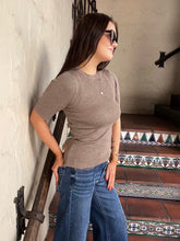 A woman wearing a fitted taupe short-sleeve knit top and blue jeans stands on a tiled staircase indoors. She is also wearing dark sunglasses and a delicate necklace.