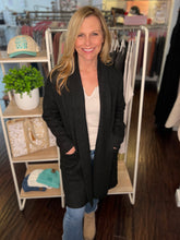 Woman standing in a clothing store wearing a black cardigan and smiling.