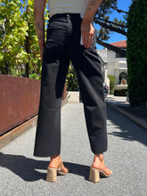 A person shown from the back wearing loose black wide-leg cropped pants, a white top, and beige block-heel mule sandals on an outdoor paved walkway with greenery and a house in the background.
