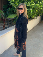 A woman wearing a long black faux fur coat with a rust-brown floral pattern, dark pants, and sunglasses, standing outdoors on a concrete walkway next to a white planter box and green foliage.