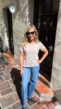 A woman wearing a light beige short-sleeve t-shirt and blue jeans stands on a brick patio in front of a building with a stucco wall and dark window frames.