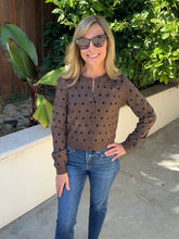 A woman wearing a brown long-sleeve button-up cardigan with black polka dots, paired with blue jeans and sunglasses, standing outdoors in front of a white wall and green foliage.