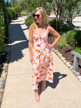 Woman in a picnic print dress standing on a sidewalk with trees and shrubs in the background