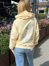 Person wearing a light yellow hoodie with an anchor design outdoors.