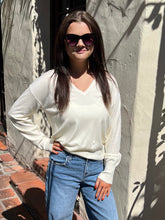 A woman wearing a cream-colored long-sleeve knit sweater and blue jeans, standing outdoors against a textured stucco wall with leaf shadows.