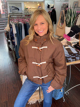 A smiling blonde woman wearing a brown puffer jacket with cream toggle closures and blue jeans, posing inside a clothing boutique.