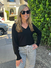 A woman wearing a black long-sleeve V-neck top, leopard print drawstring pants, and black sunglasses, standing outdoors in front of a green hedge.