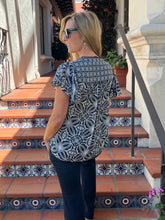 A woman wearing a black and white patterned short-sleeve blouse and dark pants, standing on outdoor tiled steps.
