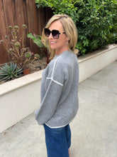 A woman wearing a gray knit sweater with white contrast stitching and blue jeans, standing outdoors on a concrete patio with green plants and a wooden fence in the background.