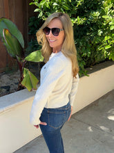 A woman with blonde hair and sunglasses wearing a cream cable-knit sweater and blue jeans, standing outdoors in front of a green hedge and a beige wall.